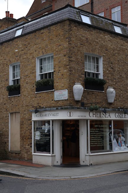 Exterior view of a three-story brick building on a street corner, featuring large display windows with white framing on the ground floor, housing Chelsea Green laundry and dry cleaning services. The b