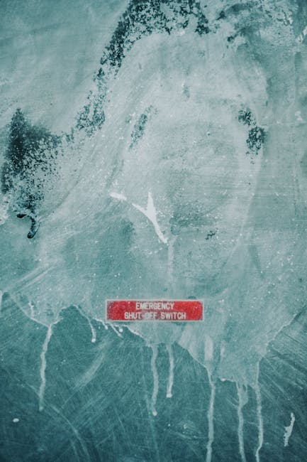 A close-up of a weathered, teal-painted wall with visible scratches, peeling paint, and stains. In the lower center, there is a red rectangular emergency shut-off switch with white text reading 'EMERGENCY SHUT-OFF SWITCH.' The surface shows signs of wear and some paint drips, indicating a need for cleaning or maintenance. The lighting highlights the texture of the wall, and the image emphasizes the importance of surface cleaning and upkeep in managing safety and hygiene in commercial spaces. This image relates to [PAGE_TITLE] by illustrating the condition of surfaces that may require deep cleaning or sanitisation. The setting appears to be an industrial or utility area, and the wall's condition underscores the significance of regular cleaning for safety compliance, as provided by Putney Carpet Cleaning.