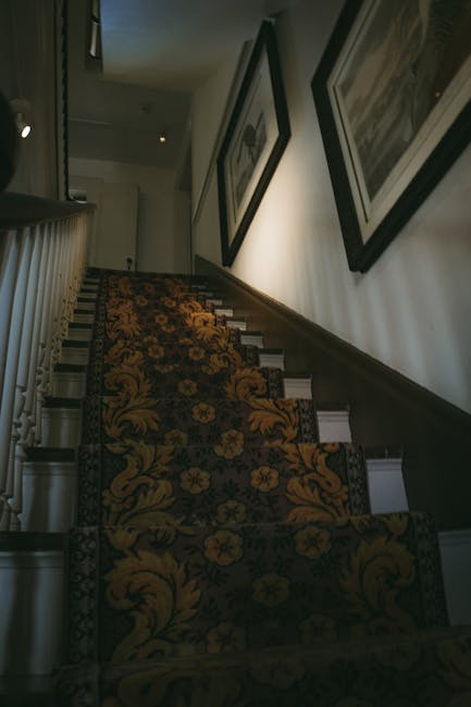 Interior view of a staircase in a Victorian home, featuring a decorative floral-patterned carpet runner in shades of gold, brown, and black, which covers wooden steps leading upward. The staircase is illuminated by soft, natural light filtering from an unseen window, highlighting the clean and well-maintained surface. The wall adjacent to the stairs displays three framed black-and-white photographs or artworks, evenly spaced and hung at eye level. A white wooden handrail runs along the left side of the staircase, adding a traditional touch to the setting. This image reflects the type of surface cleaning and maintenance services offered by Putney Carpet Cleaning, emphasizing attention to detail in domestic cleaning within elegant Victorian homes.