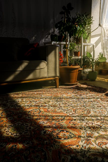A sunlit Victorian living room featuring a traditional patterned rug with intricate floral motifs in shades of orange, cream, and navy covering a wooden floor. On the left, a beige sofa with wooden legs is positioned against a dark wall, casting a shadow across part of the rug. Near the window, several potted houseplants of various sizes and types are arranged on a white metal plant stand and on the floor, receiving natural light that highlights their green foliage. The sunlight creates contrasting light and shadow areas on the rug and floor, emphasizing the cleanliness and well-maintained appearance of the room. The scene depicts a tidy, inviting domestic space, consistent with surface cleaning and maintenance routines provided by Putney Carpet Cleaning.
