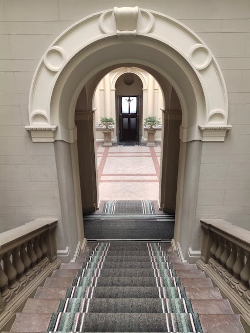 Interior view of a staircase in a Victorian home, featuring a decorative floral-patterned carpet runner in shades of gold, brown, and black, which covers wooden steps leading upward. The staircase is illuminated by soft, natural light filtering from an unseen window, highlighting the clean and well-maintained surface. The wall adjacent to the stairs displays three framed black-and-white photographs or artworks, evenly spaced and hung at eye level. A white wooden handrail runs along the left side of the staircase, adding a traditional touch to the setting. This image reflects the type of surface cleaning and maintenance services offered by Putney Carpet Cleaning, emphasizing attention to detail in domestic cleaning within elegant Victorian homes.