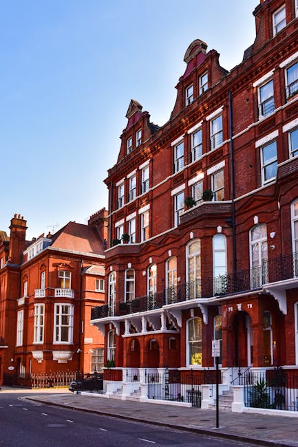 A street scene featuring a row of Victorian-style residential buildings with red brick facades, white decorative trims, and arched windows, situated along a paved road with parked cars on the side. Th