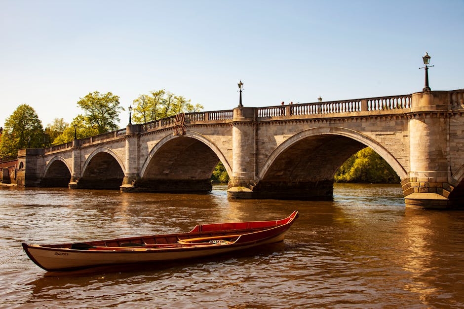 A stone river bridge with multiple arches, featuring decorative lampposts along the balustrade, spans over a calm river. Below, a small wooden boat with a red interior is floating on the water. The scene is illuminated by warm sunlight, highlighting green trees lining the riverbank in the background. The image captures a serene riverside setting associated with Putney, as featured on the Putney Bridge upholstery cleaning page of putneycarpetcleaning.com, emphasizing the importance of surface cleaning and maintenance to preserve the scenic environment surrounding riverside flats.
