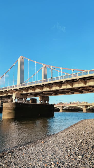 A stone river bridge with multiple arches, featuring decorative lampposts along the balustrade, spans over a calm river. Below, a small wooden boat with a red interior is floating on the water. The scene is illuminated by warm sunlight, highlighting green trees lining the riverbank in the background. The image captures a serene riverside setting associated with Putney, as featured on the Putney Bridge upholstery cleaning page of putneycarpetcleaning.com, emphasizing the importance of surface cleaning and maintenance to preserve the scenic environment surrounding riverside flats.